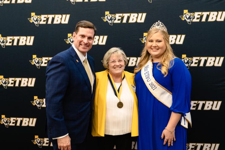 Jeanne Beth (Gibbs) Gray (center) is joined by President Dr. Blair Blackburn (left) and Kathryn Pedroza, the 65th Miss ETBU. Two women and a man smiling at the camera in front of a navy blue ETBU backdrop