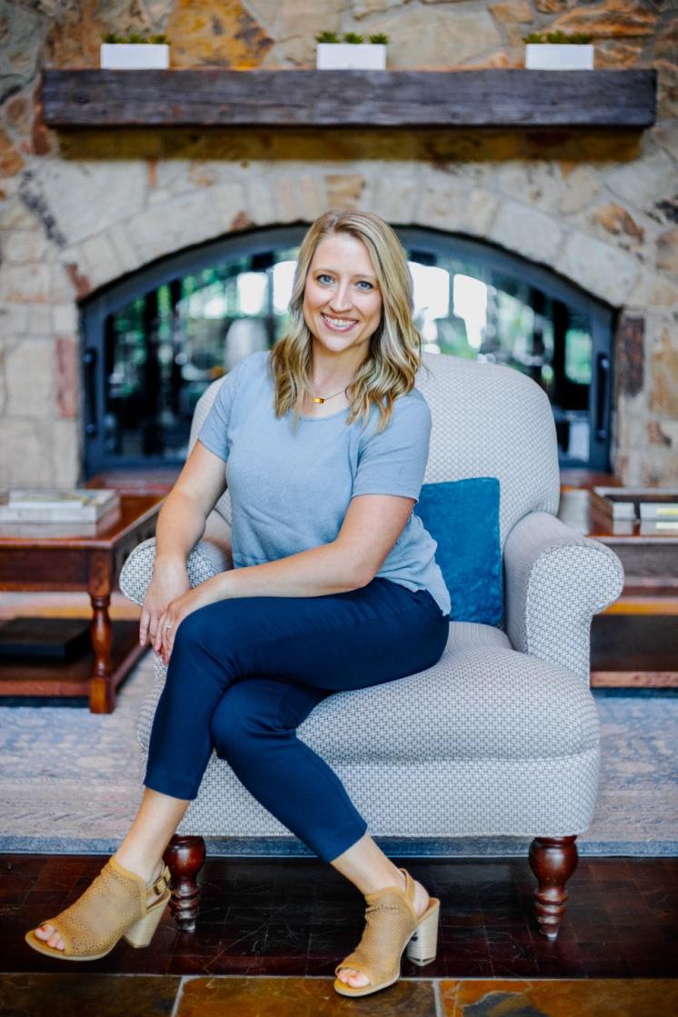 A woman in a blue shirt and jeans sitting in a grey chair in front of a stone fireplace.