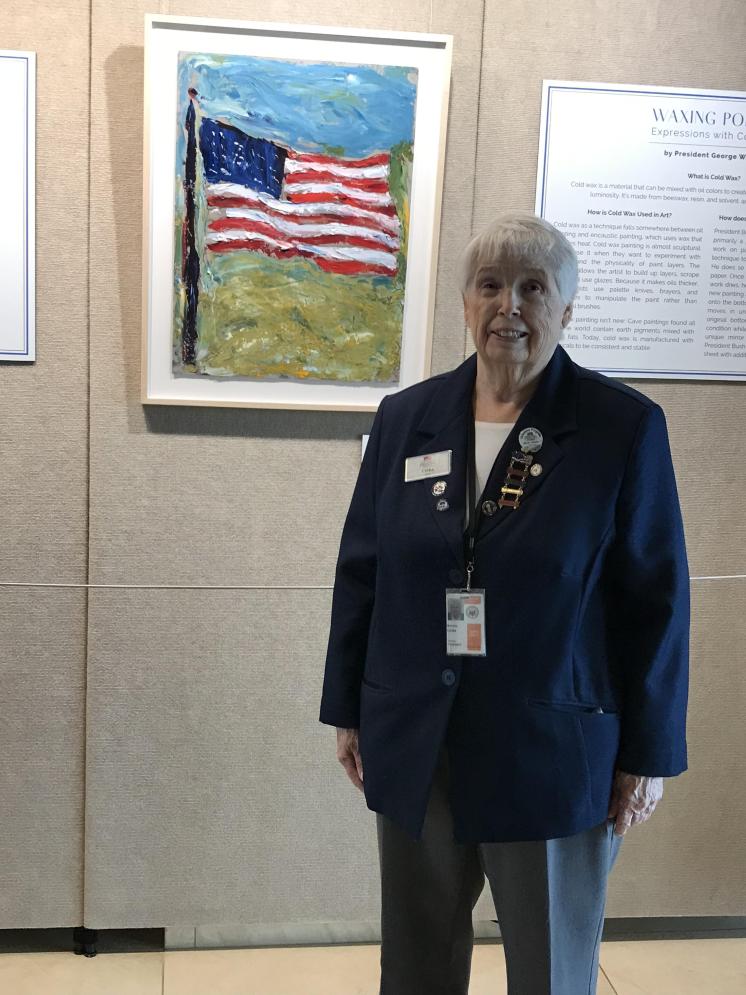 An older woman in a navy blazer in front of a painting of an American flag displayed on a beige wall.