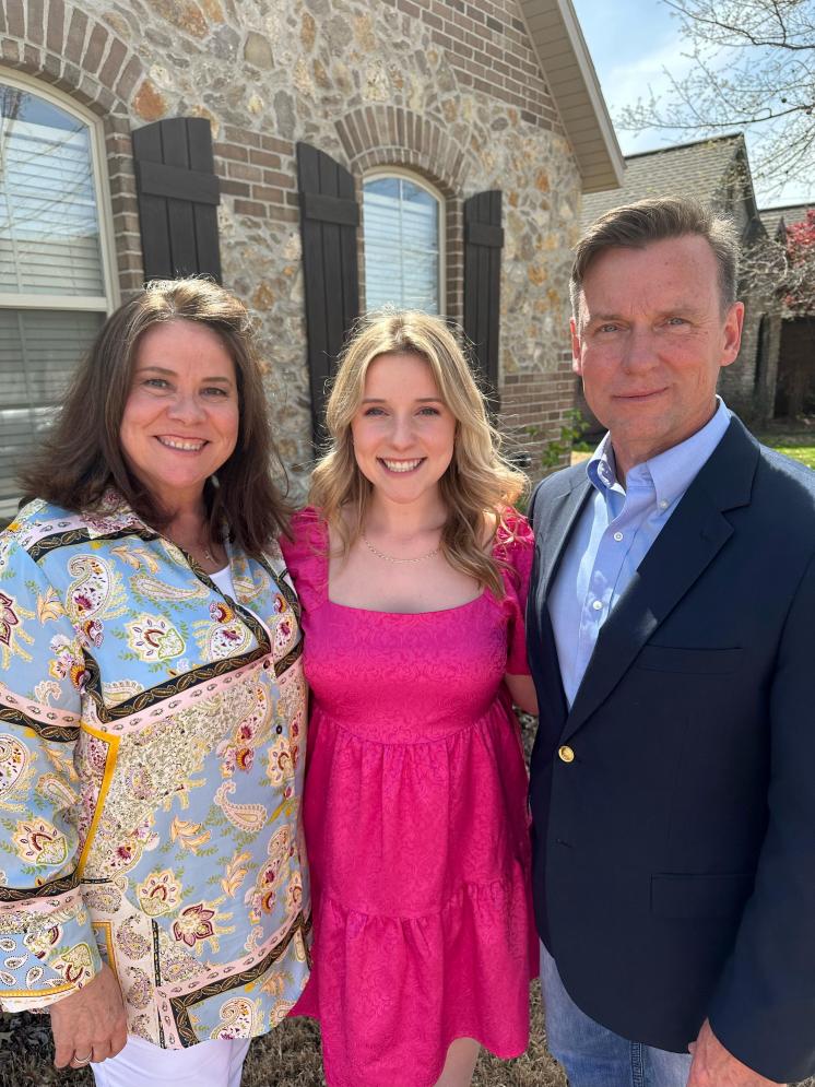 This image shows three people standing together in front of a brick and stone house with arched windows and dark wooden shutters. The woman on the left is wearing a light blue patterned blouse with white pants. The young woman in the center is wearing a bright pink dress with short sleeves and a square neckline. The man on the right is dressed in a dark navy blazer, light blue dress shirt, and jeans. All three are smiling and standing close together, posing for the photo in a sunny outdoor setting.