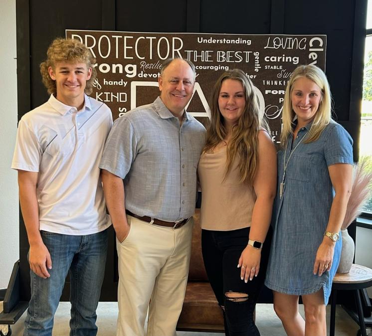 A family photo featuring four people standing side by side, smiling at the camera. From left to right: a young man in a white polo shirt and jeans with curly hair, an older man in a light gray short-sleeved button-up shirt and beige pants, a young woman in a beige sleeveless top and black ripped jeans with long hair, and a woman in a denim dress with blond hair. Behind them is a brown sign with various positive words, including "PROTECTOR," "STRONG," "THE BEST," and "LOVING."