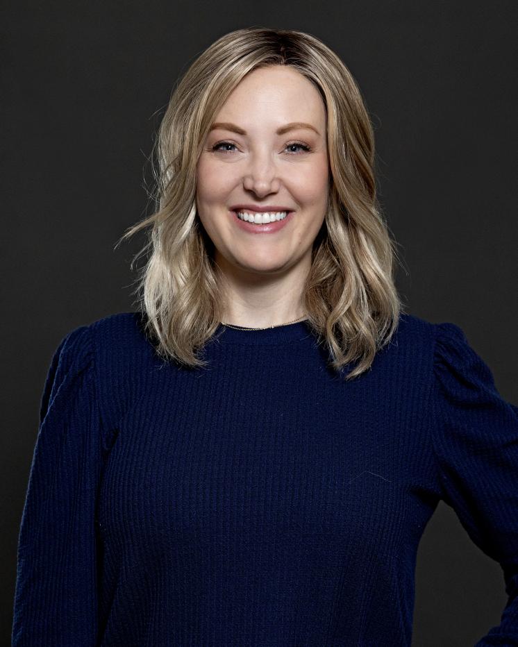 A smiling woman with shoulder-length blonde hair is wearing a dark blue long-sleeved shirt, posed against a plain dark background.