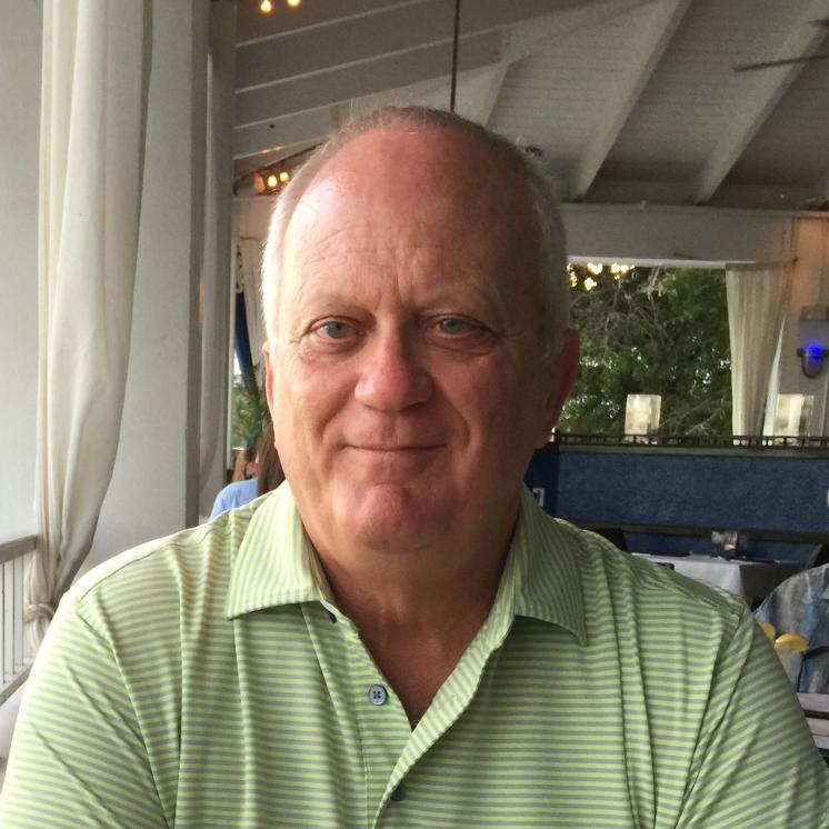 A man with short, light hair is sitting at a table in a restaurant, wearing a light green striped polo shirt, with a calm expression. The restaurant has a casual, open-air design with white curtains and ceiling fans.