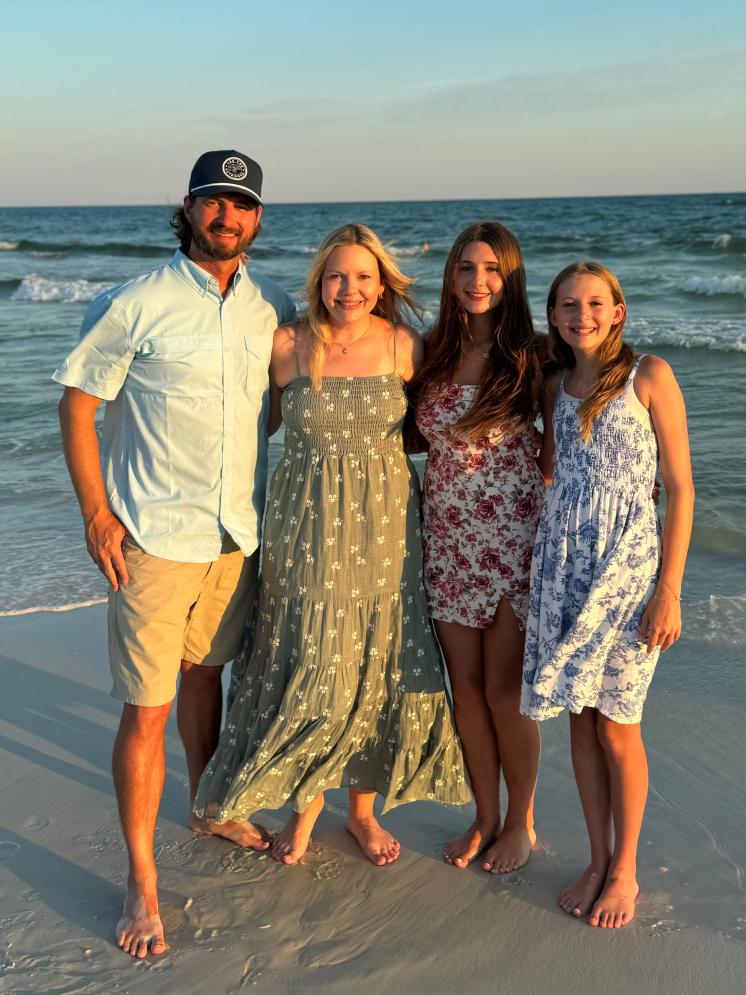 Family of four, Dad mom and two daughters. Daughters wearing sundresses on the beach while the dad wear shorts and a hat.