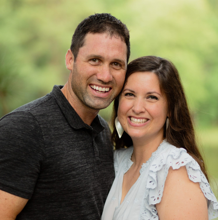 A smiling couple standing closely together outdoors with a green, blurred background.