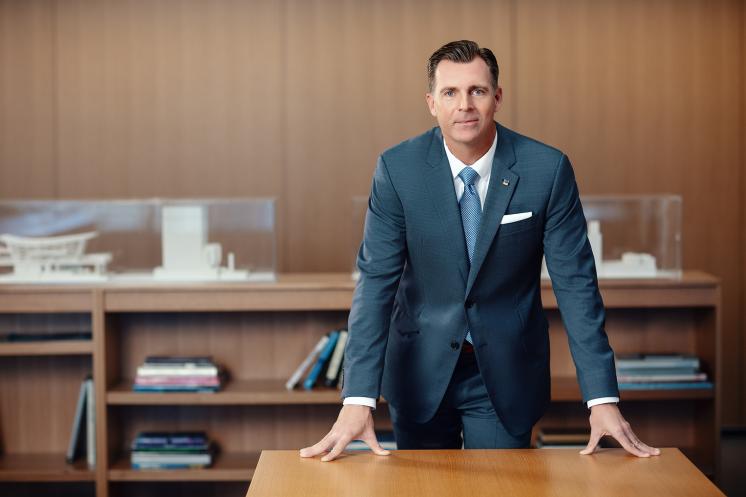 Man in a suit standing beind a desk leaning on it.