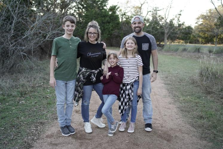 A family of five posing outdoors on a dirt path, surrounded by trees.
