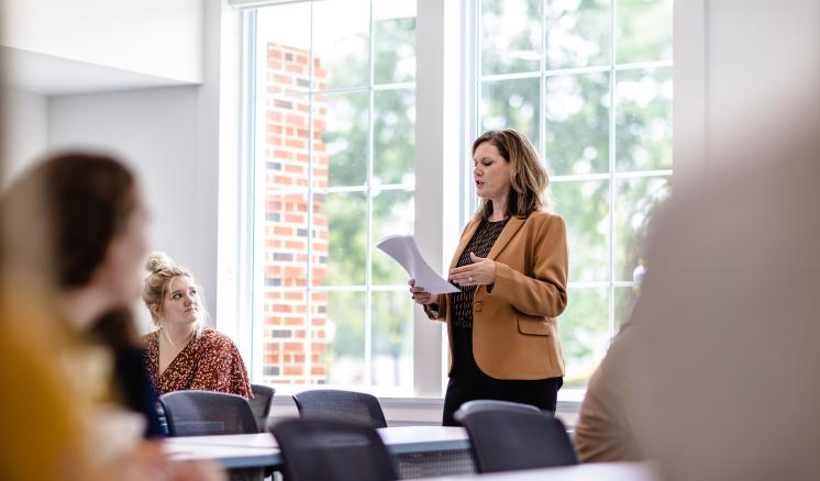 School of Education Full Accreditation Woman standing and presenting in a classroom with seated attendees and large windows in the background.