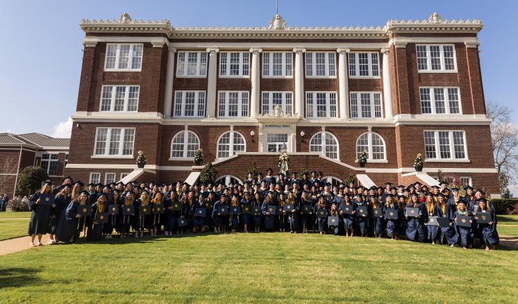 Fall 2023 Commencement Large group of graduates posed with diplomas in front of brick academic building smiling at the camera