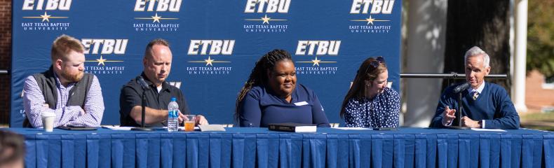Men and women sitting at a table with a blue tablecloth in front of a blue ETBU backdrop outside