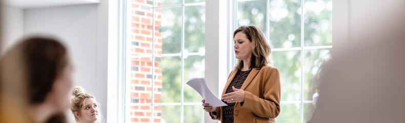  Woman standing and presenting in a classroom with seated attendees and large windows in the background.