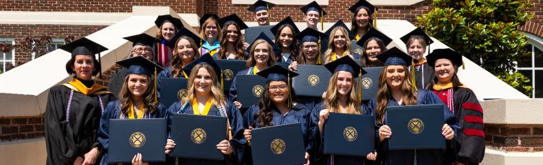 A group of graduates in caps and gowns stand on steps outside a building, holding their diplomas. They are smiling alongside faculty members, celebrating their graduation.