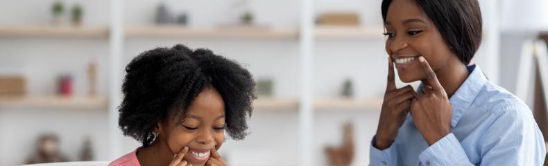 A child and speech-language pathologist sit together at a table, both smiling warmly and pointing to their cheeks in front of a small mirror.